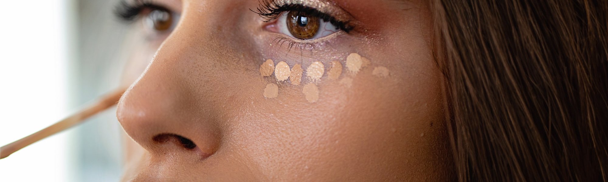 Junge, geschminkte Frau trägt unter ihren Augen Concealer in Tupfenform auf.